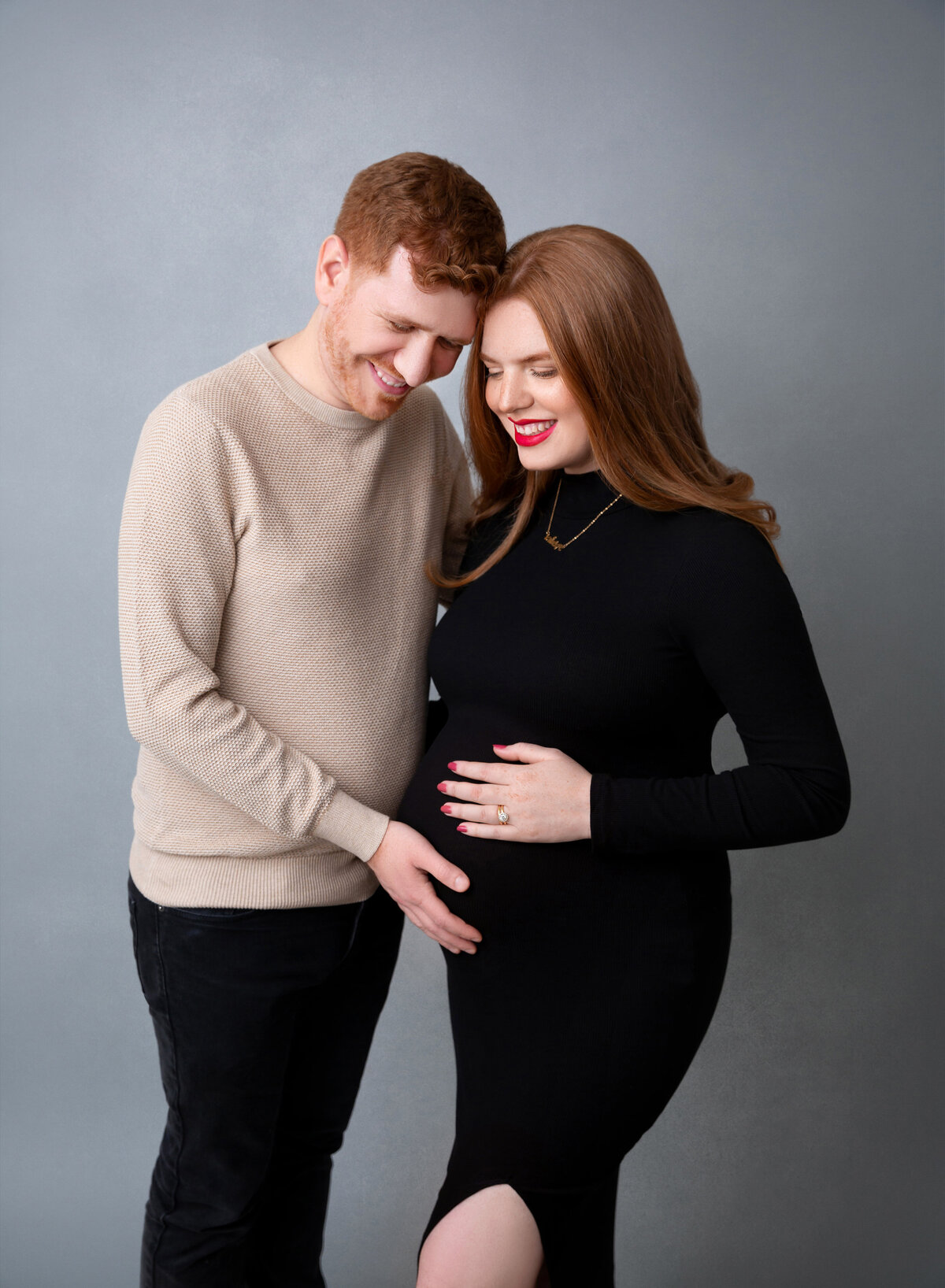 Expectant mom and dad pose for maternity photos in Brooklyn, NY. Mom and dad are standing shoulder-to-shoulder with their heads together and touching the baby bump.