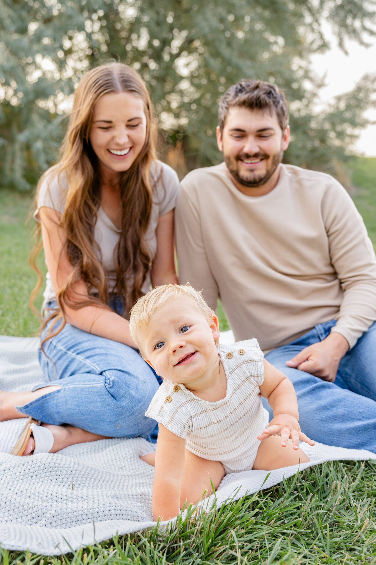 A baby with bright blue eyes crawls toward the camera while his parents laugh in the background, enjoying a peaceful family outing in Eagle Mountain, Utah.