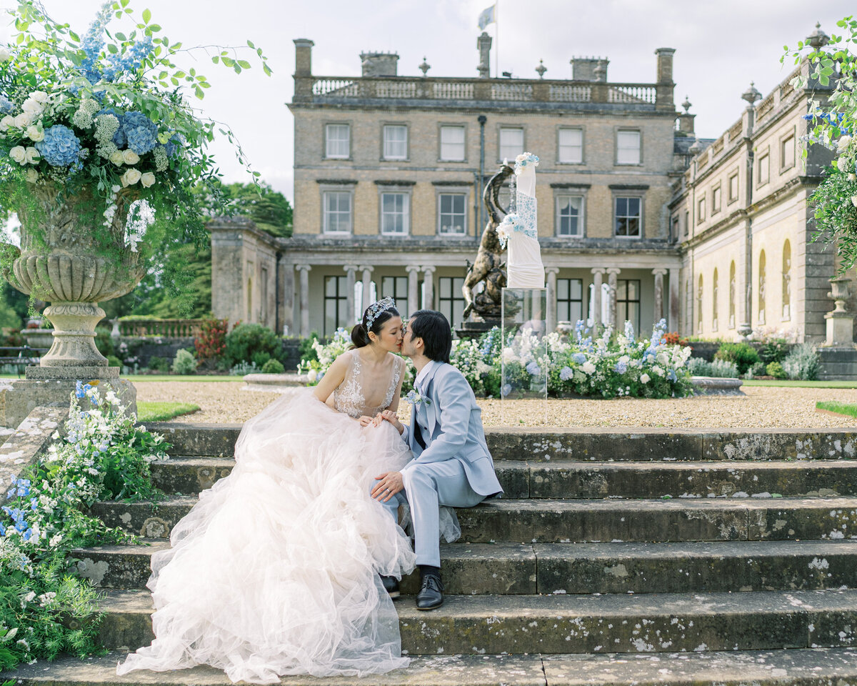Bride and groom kissing on the steps of Somerley House wedding venue, surrounded by white and blue florals