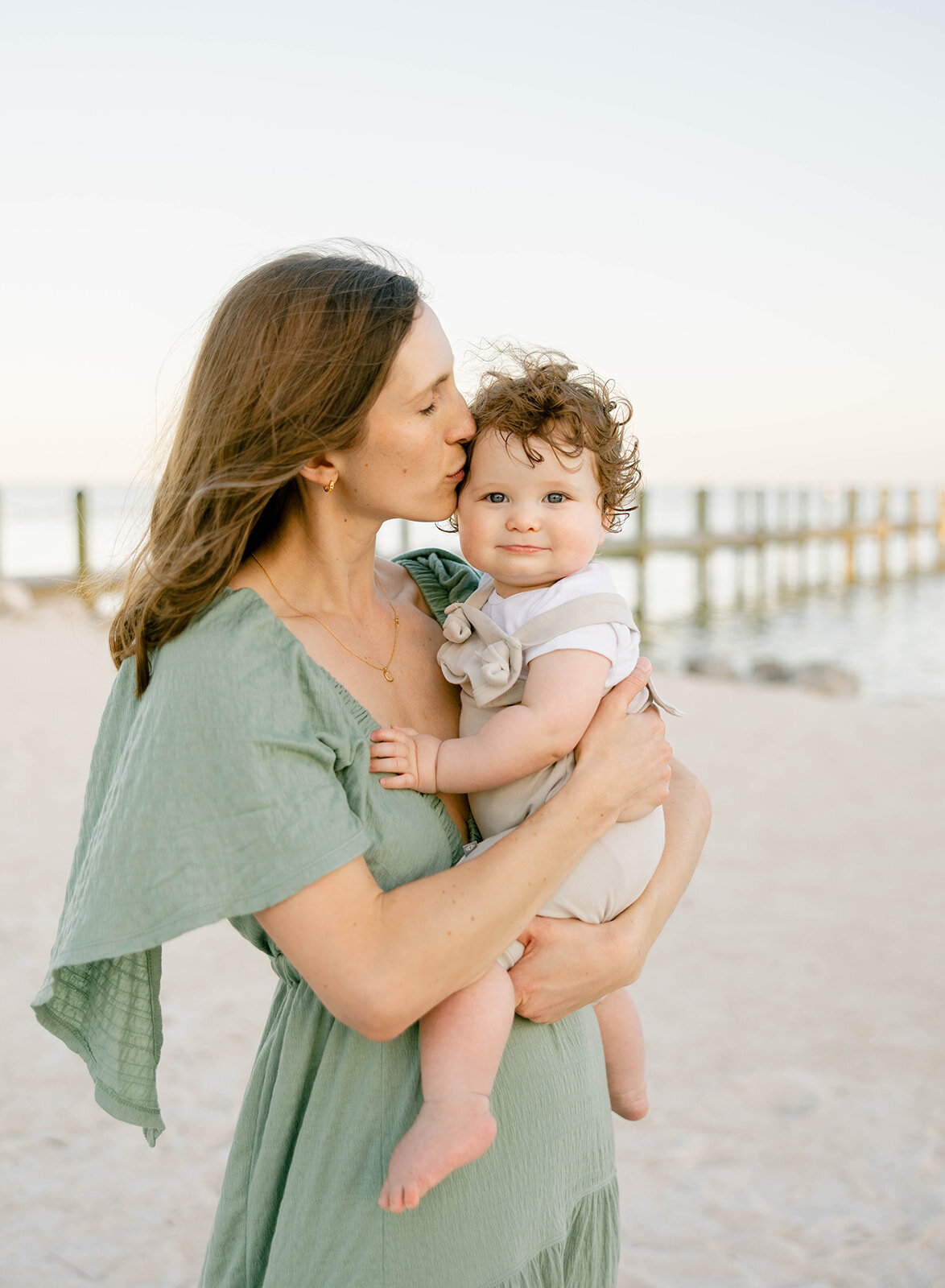 A mother kisses her curly-haired baby while standing on the sandy shore, with a wooden pier in the background. A heartfelt moment captured during a Florida Keys family session.