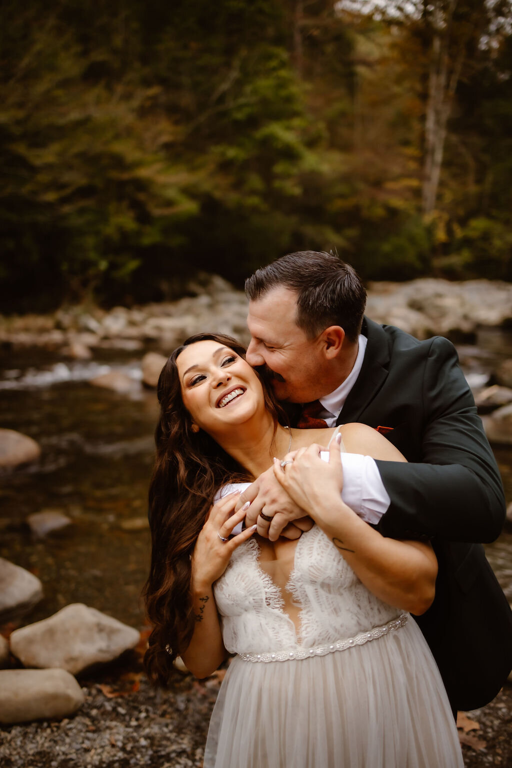 Groom embracing the bride from behind as they laugh together near the river at Greenbrier, capturing a candid moment of joy during their eloping to Gatlinburg celebration.
