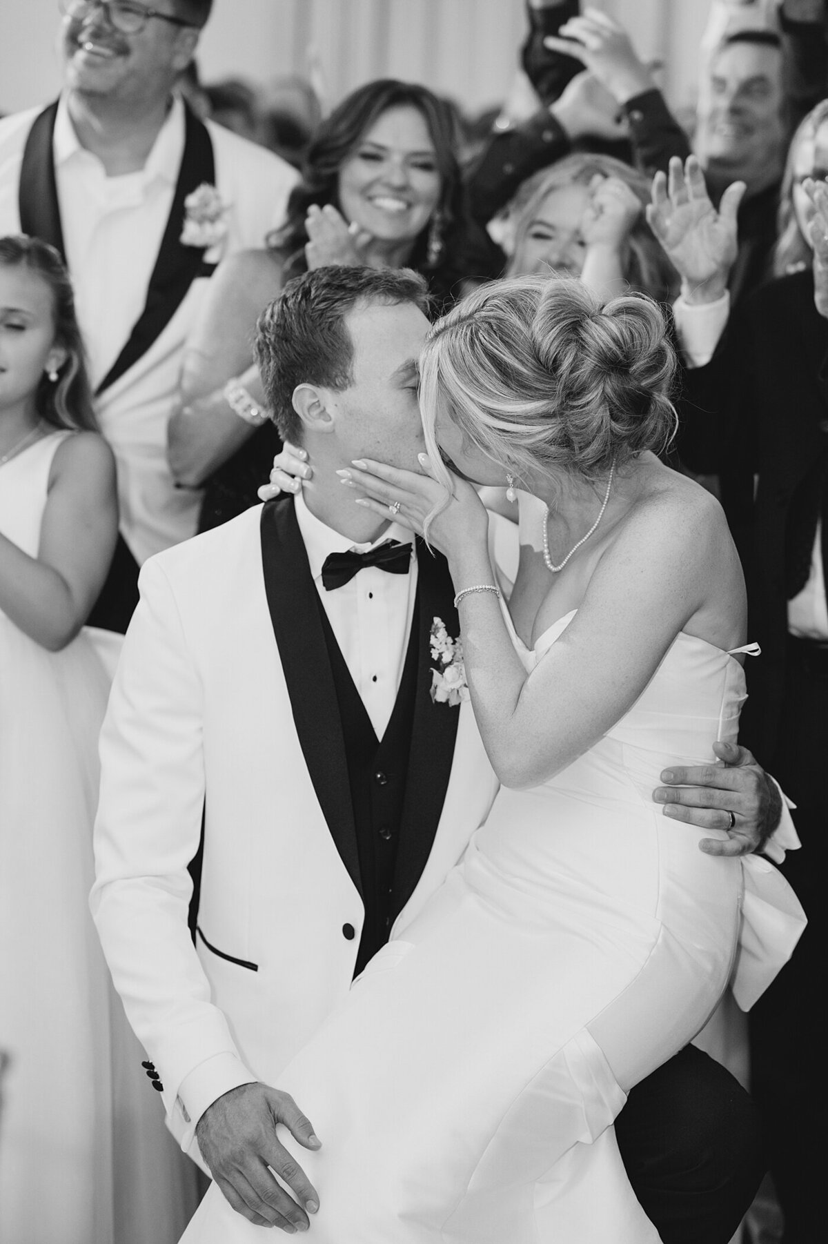 black and white wedding photo of bride sitting on her grooms lap and kissing him with their bridal party surrounding them and cheering