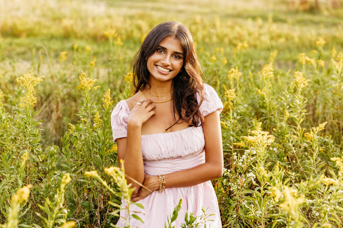 brown haired teen girl in a light pink dress kneeling by goldenrod and smiling as she rests her hand on her chest.