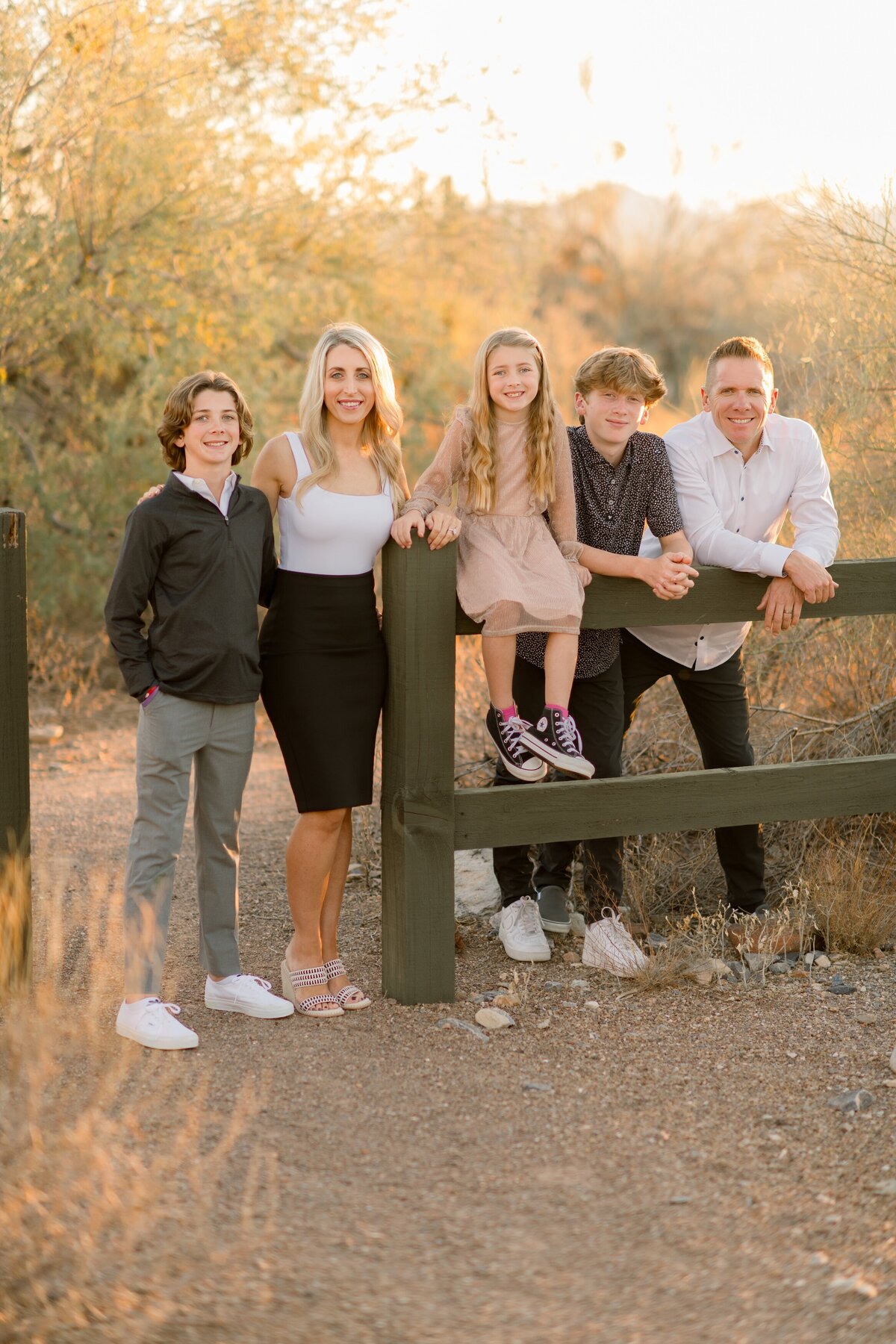 family of 5 with the mom and dad standing in the scottsdale arizona desert one little girl is sitting on the fence in a pink dress and her brothers are leaning on the fence