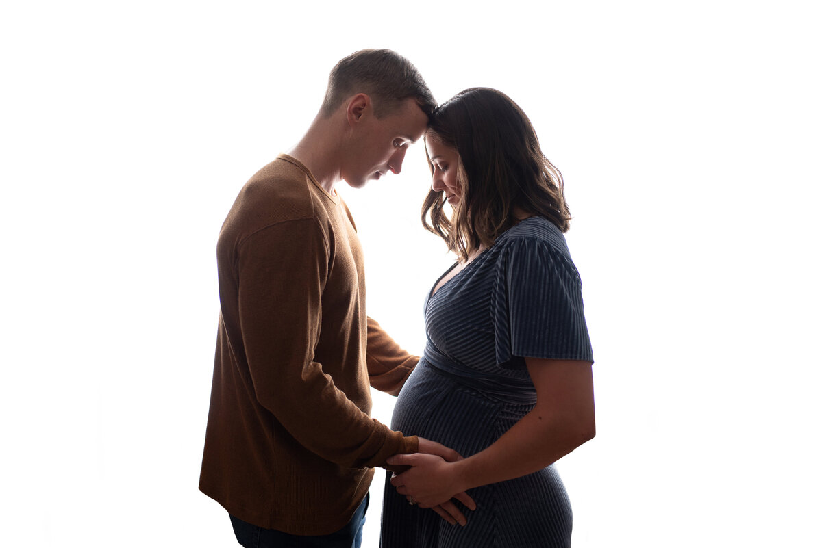 Pregnant couple at 32 weeks of pregnancy posing for maternity portrait facing each other with heads together holding woman's belly with white back light
