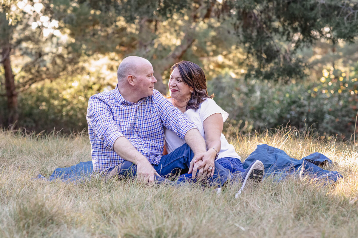 A husband and wife holding hands and sitting on a blanket in a park, looking at each other and smiling.