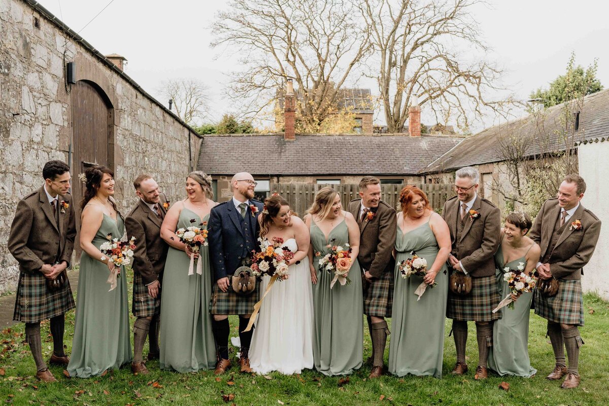 A wedding party poses outdoors in a large barn courtyard for their wedding photographer. The group includes eight people in sage green dresses and kilts, holding bouquets, with a couple at the centre sharing a joyful moment. Bare trees and rooftops rise above.