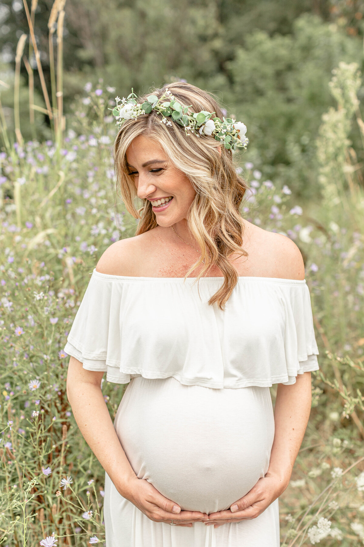 Pregnant woman in white off-the-shoulder dress holding pregnant belly and standing in a big tall grassy field with wildflowers. Portland Maternity Photographer