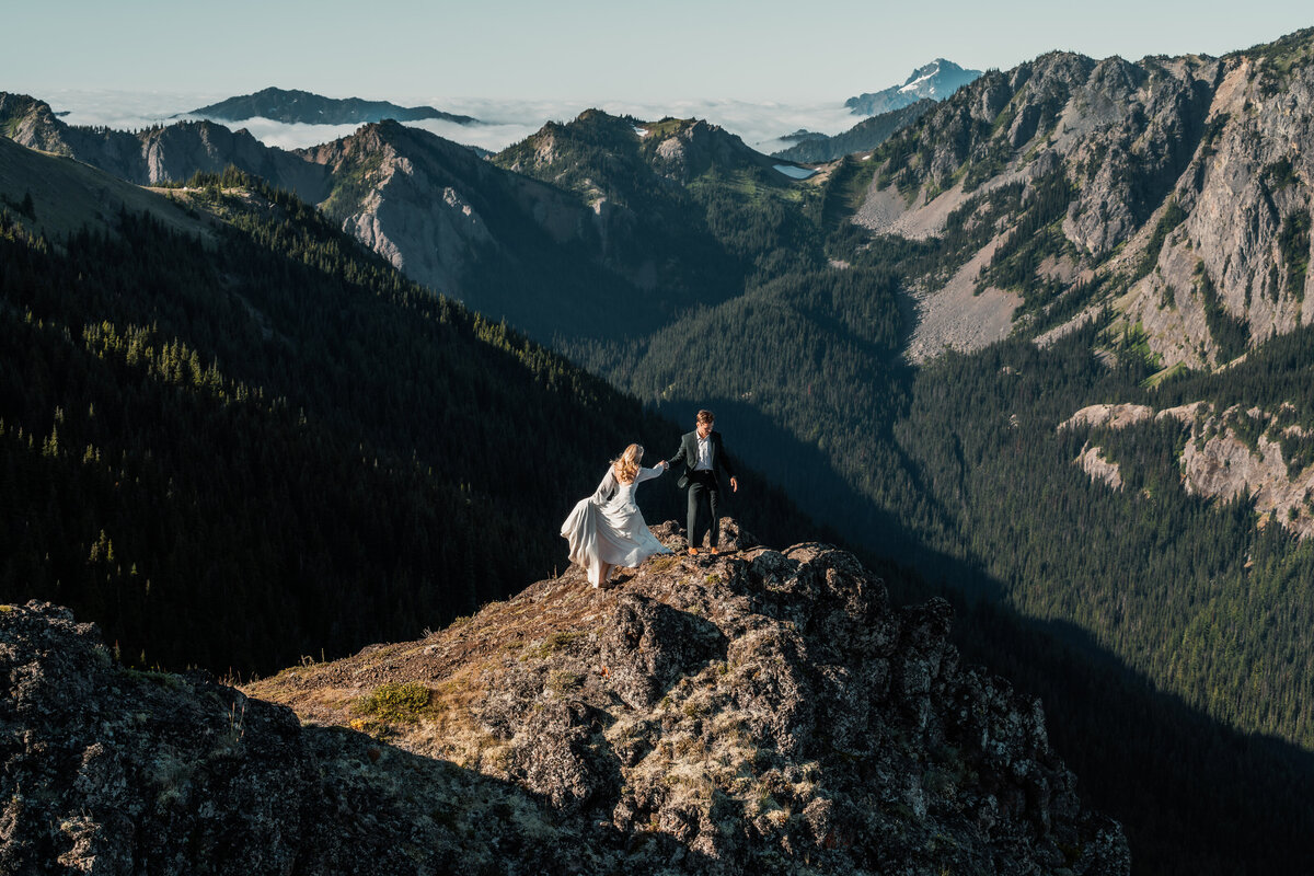 a couple in their wedding attire climb on the edges of a cliff being kissed by the sun. mountains roll behind them as their adventure elopement photographers document them exploring the landscape.