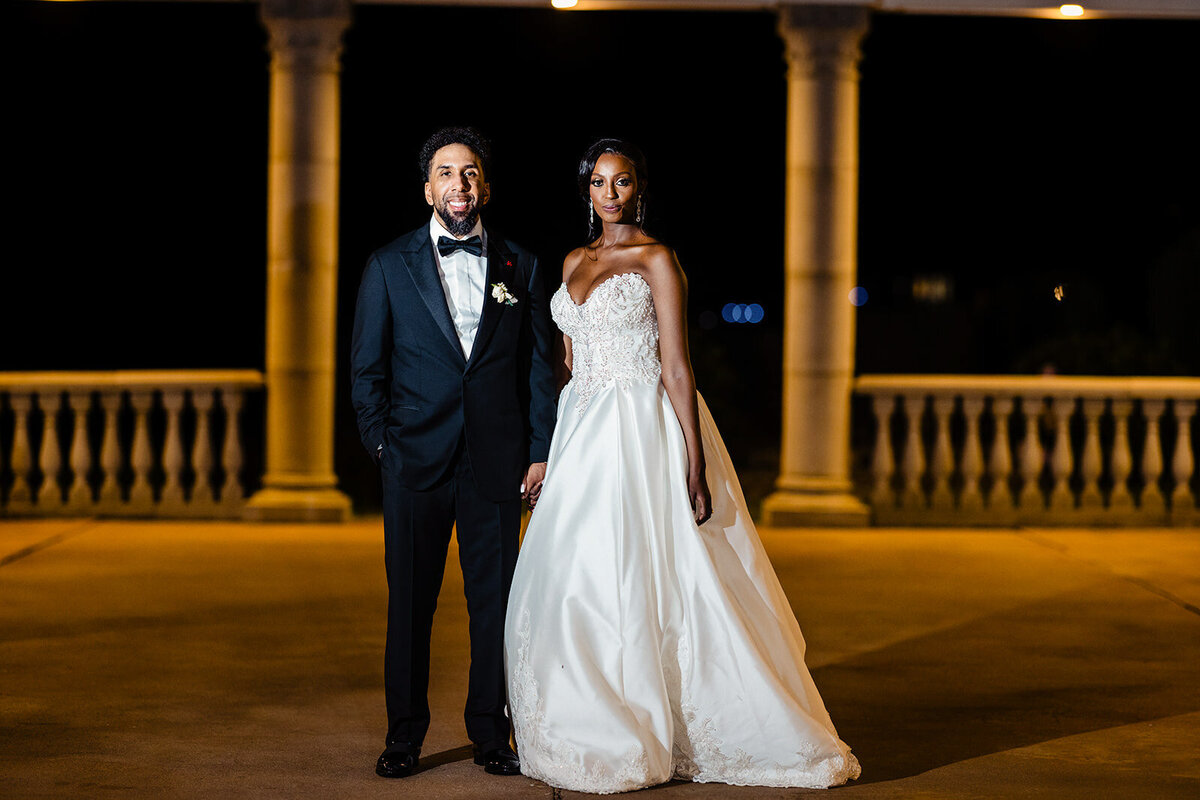 Bride and groom standing regally at night by a columned balustrade, the bride in a white dress and the groom in a black tuxedo