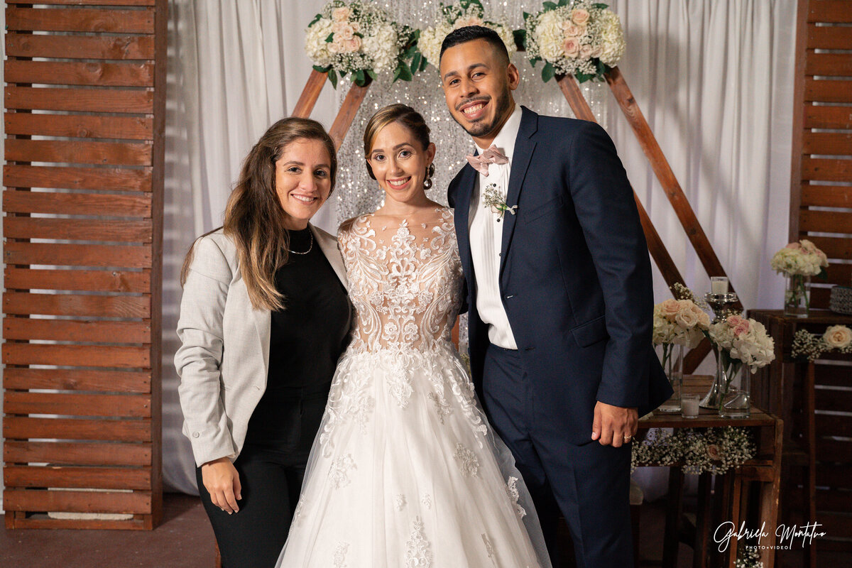 Female wedding photographer posing with a bride and groom at a floral wedding setup in San Sebastián, Puerto Rico.