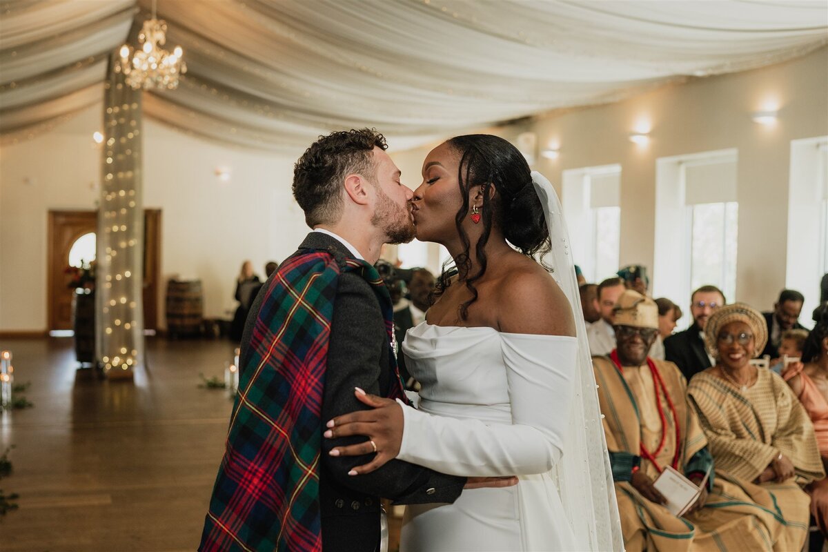 A bride and groom kiss during their wedding ceremony in a bright ballroom at Logie Country House. Guests sit in the background and applaud.