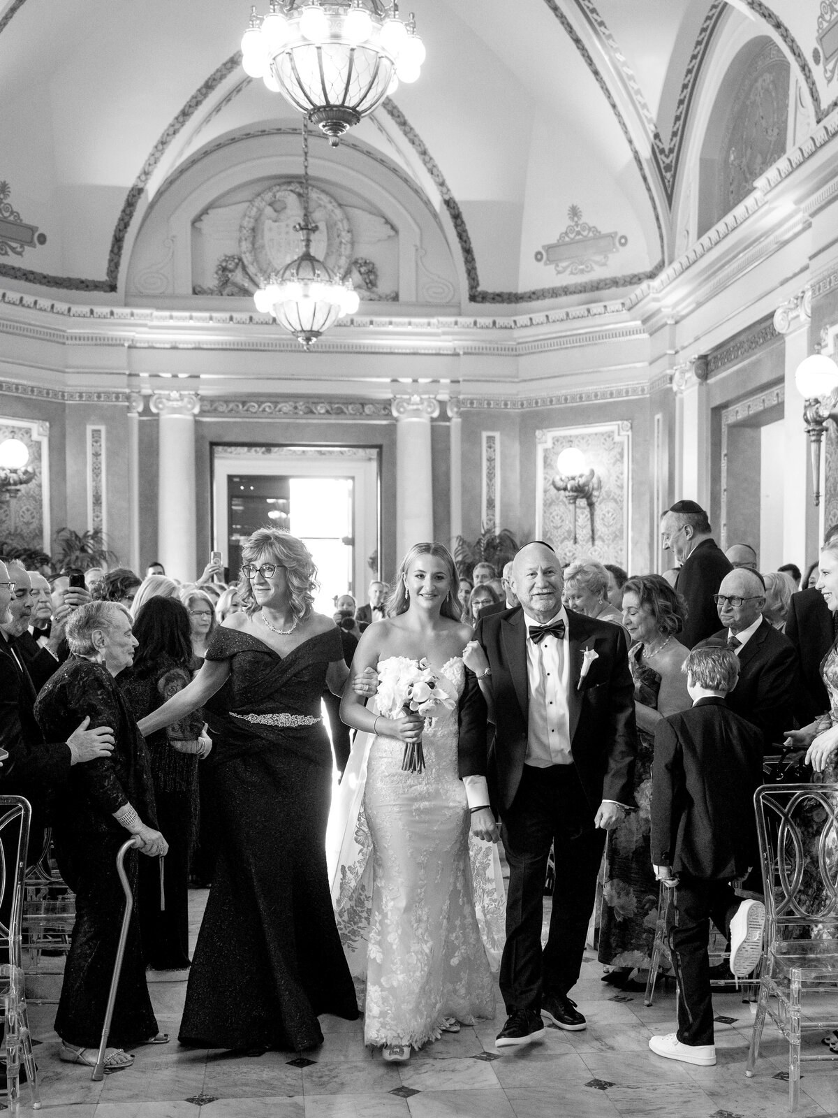 A bride in a strapless gown walks down the aisle with two companions on either side, in an ornate hall filled with guests. They are all smiling, and the guests are standing and watching. Chandeliers hang from the decorated ceiling above.