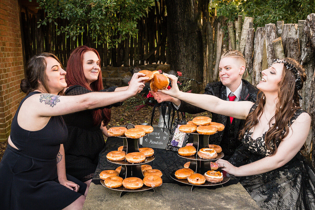 An LGBTQ+ couple on their wedding day grabs donuts from two donut towers on the table they’re at and holds them up to their persons of honor so as to “clink” donuts to say “cheers” to the couple’s successful wedding day.