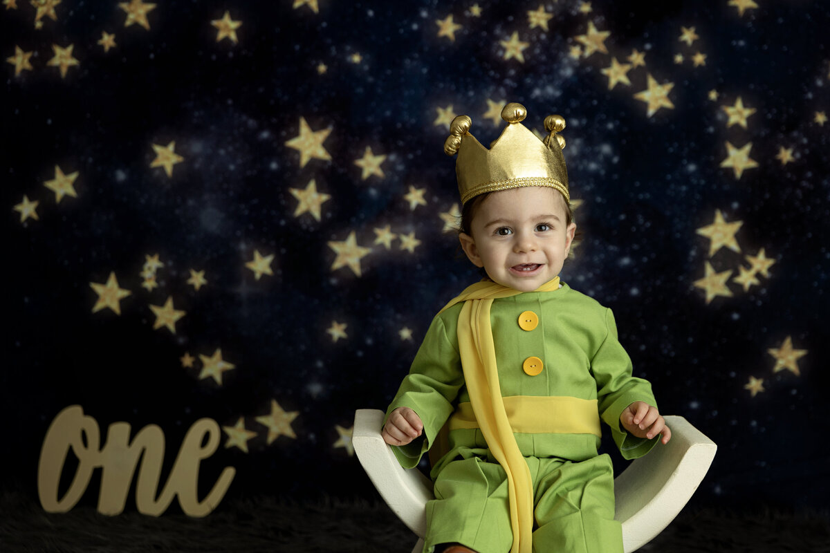 A young toddler boy dressed in a green outfit sits in a curved chair in a studio with a star backdropNew Jersey Milestone Photographer