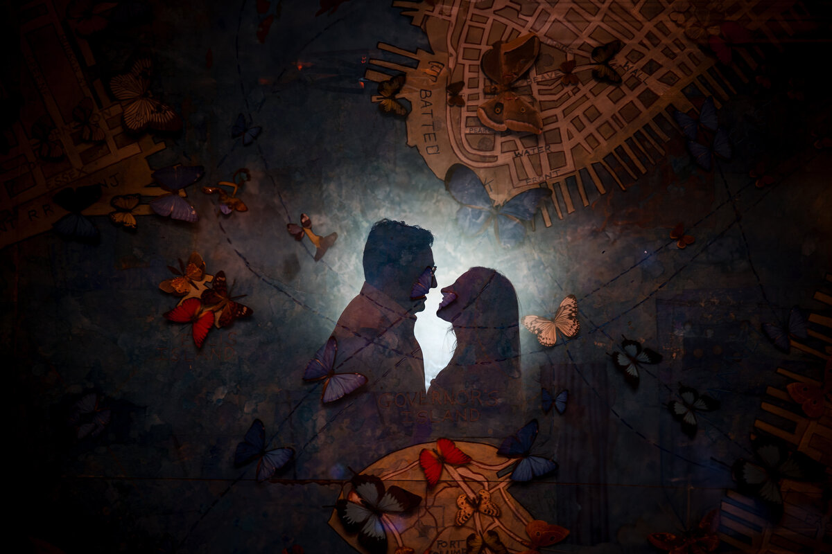 Silhouetted couple facing each other, lit from behind, overlaid on a mural map of Lower Manhattan featuring Governors Island and butterfly illustrations; the layered composition blends romance and location, with the butterflies appearing to fly across the scene.