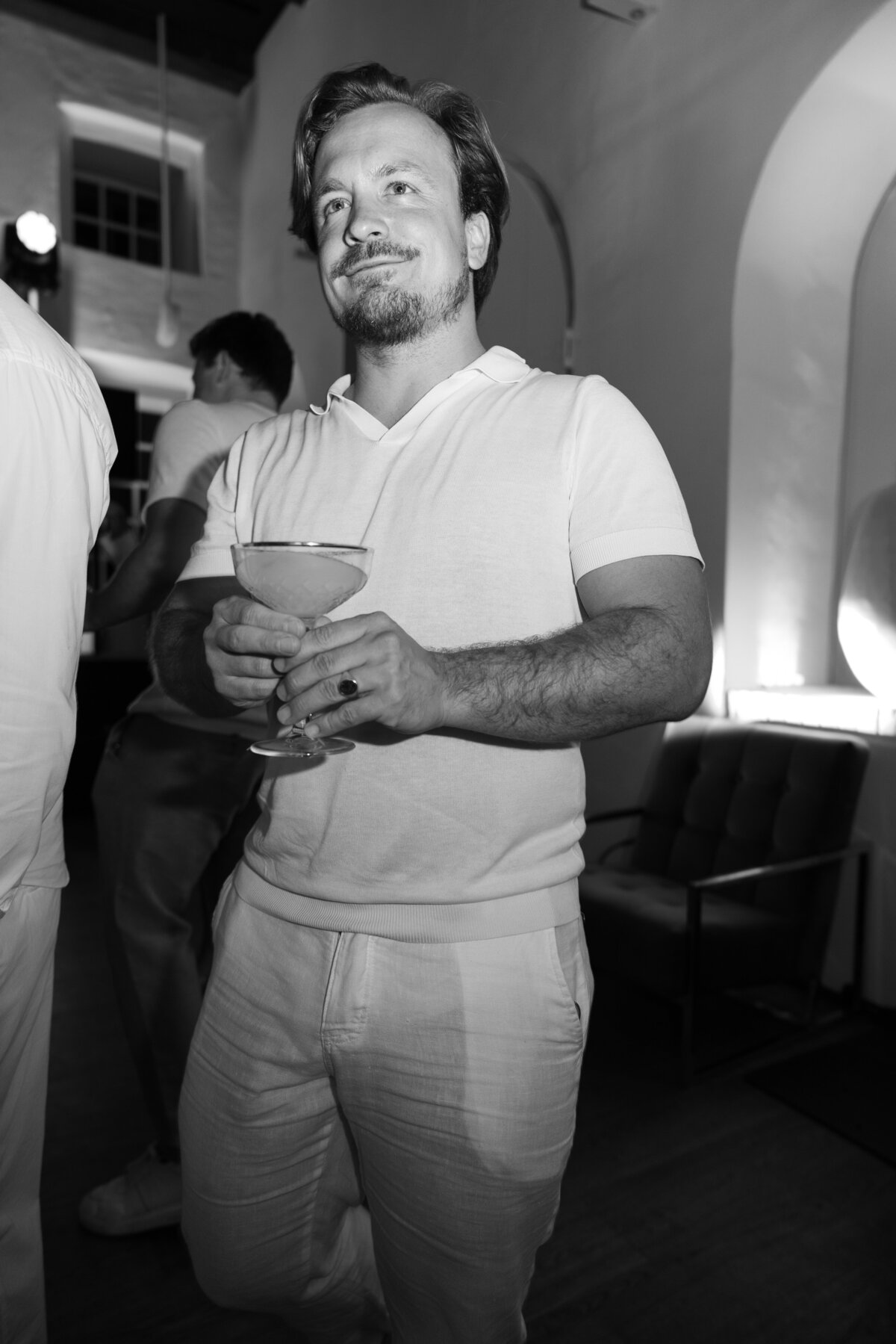 A guest enjoying a cocktail at the event, dressed in white attire that matches the elegant theme of the night. This black-and-white portrait captures the relaxed and joyful mood of the evening.