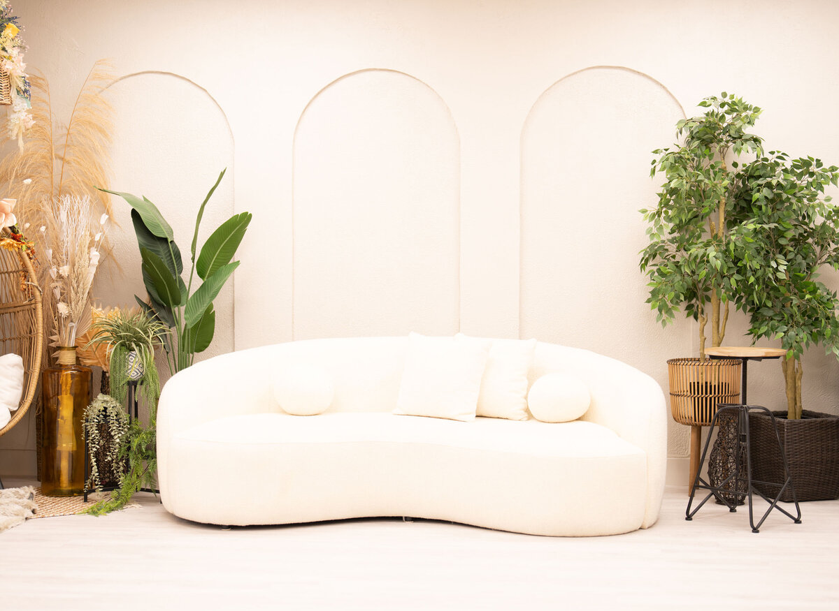 Minimalist living room with a cream curved sofa, flanked by vibrant green plants and pampas grass, set against a cream wall with subtle arch designs.