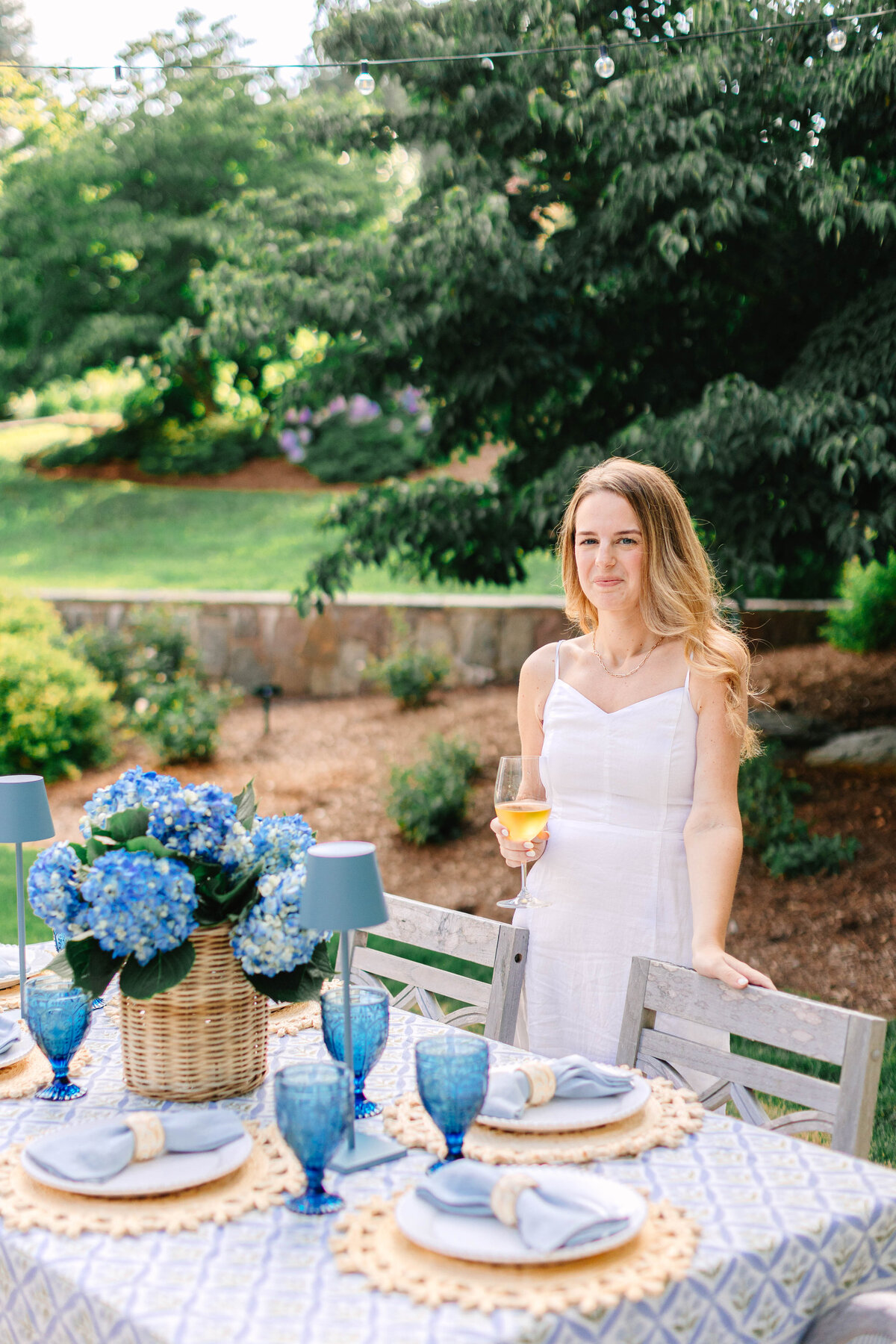 A woman in a white sundress stands beside an elegantly set outdoor dining table, holding a glass of white wine. The table features blue glassware, woven placemats, and a wicker basket filled with blue hydrangeas, set against a lush garden backdrop with string lights overhead