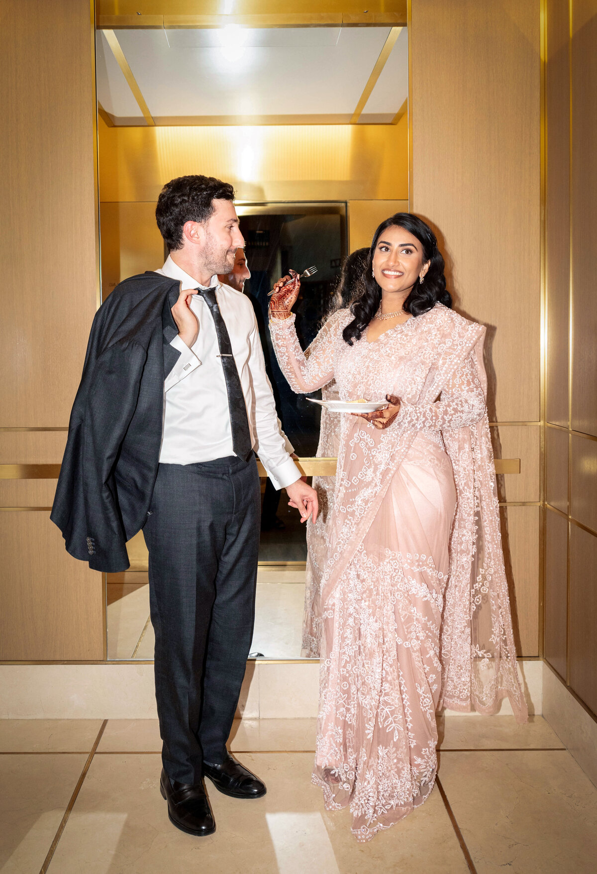 A man in a suit holds his jacket over his shoulder, standing beside a woman dressed in an elegant pink sari with lace details. They are smiling and appear to be enjoying a lighthearted moment in a brightly lit hallway.