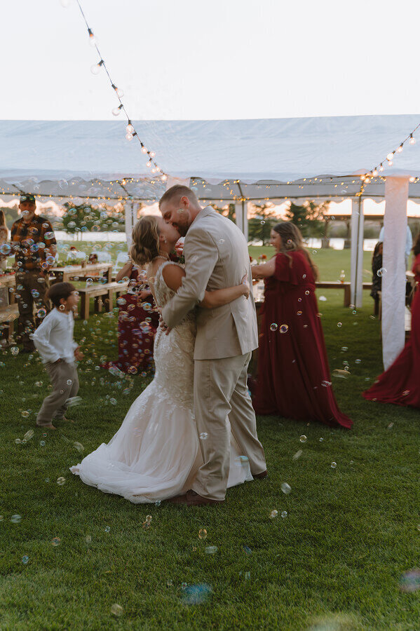 A bride and groom kiss at their wedding reception. Behind them are wedding guests and all around them are bubbles.
