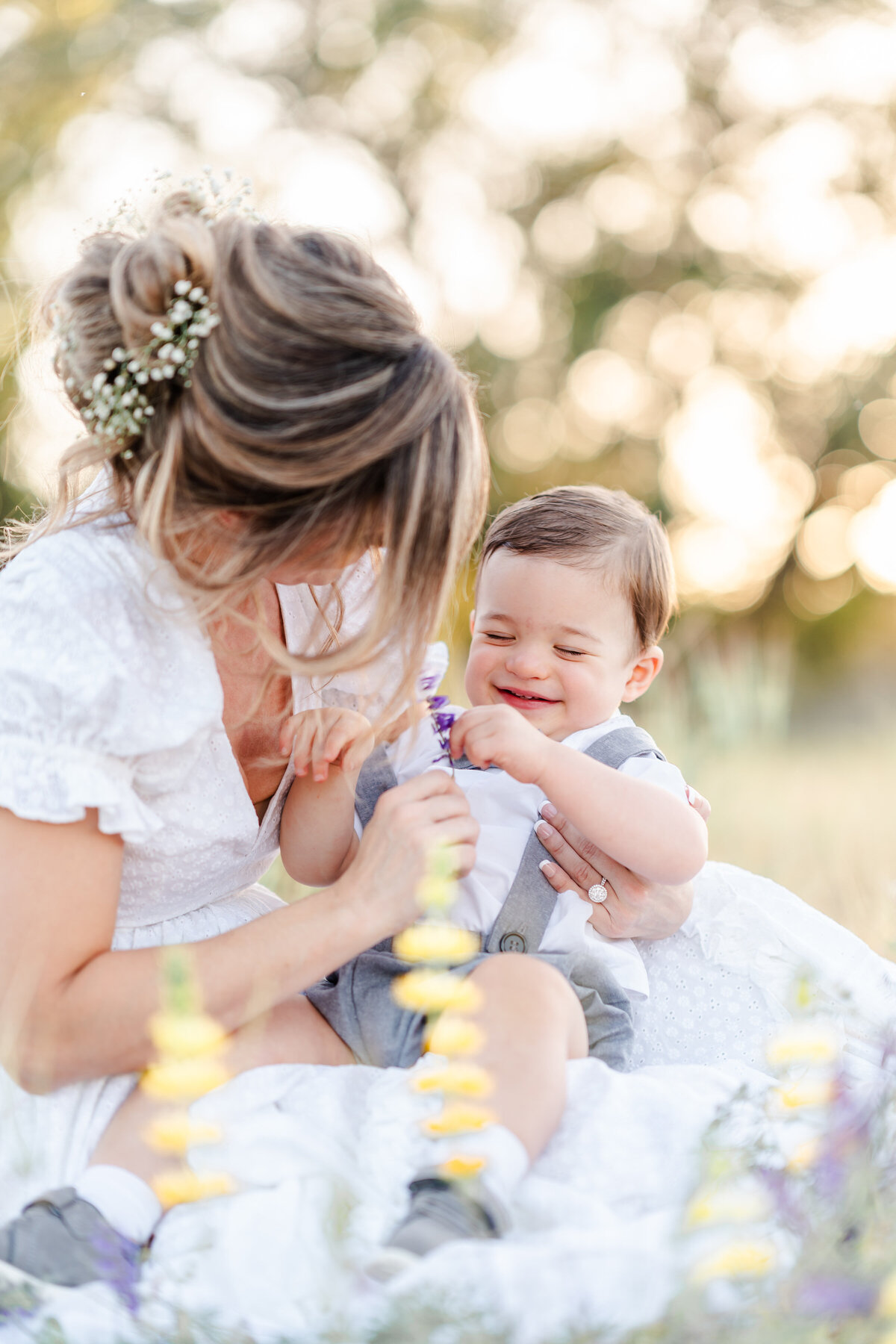 A mother wearing a white gown with a floral headpiece of babies breath sits in an open field of flowers with her toddler son in grey overalls while he smiles and holds a flower photographed by Bay Area Family Photographer, Light Livin Photography.