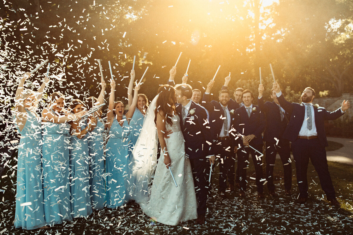 Wedding  party pops confetti in the sunlight as bride and groom kiss.