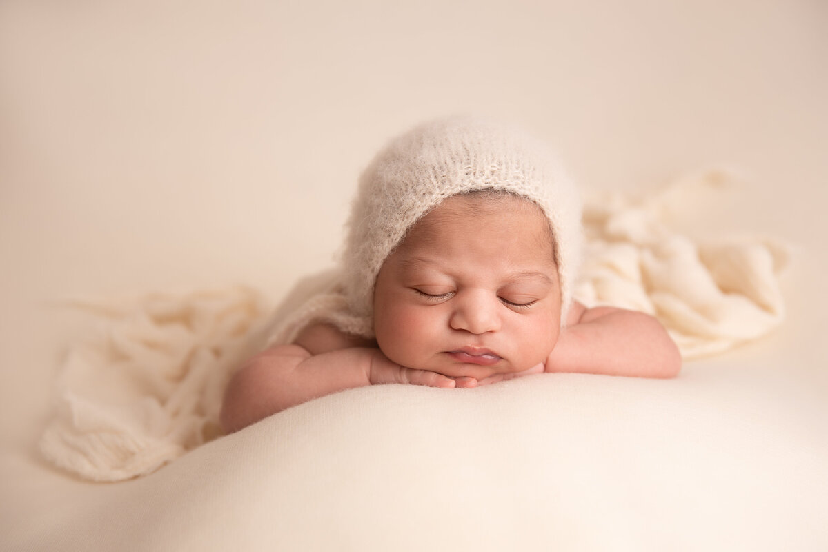 Newborn baby girl wearing a cream knitted bonnet sleeping on her tummy on a cream fabric background.