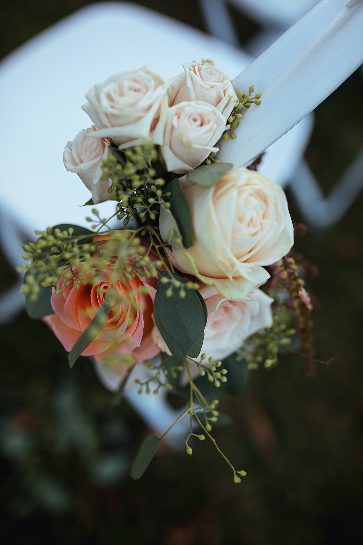 Detail photo of chair marker flowers for a wedding ceremony at Cheekwood. The white garden chair has seeded eucalyptus, peach and ivory roses tied to the side.