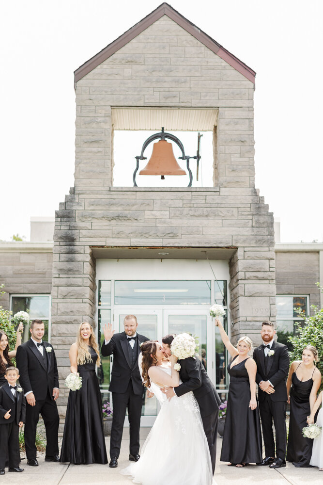 bride and groom kissing in front of a bel tower as bridesmaids and groomsmen cheer