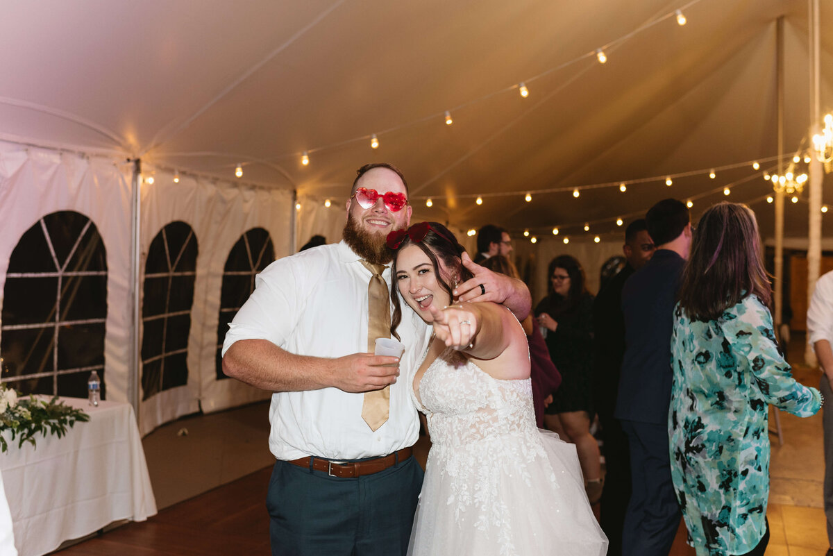 bride and groom at their wedding reception with groom in heart shaped glasses and the bride pointing to the camera in an outdoor tent decorated by string lights captured by Charlottesville wedding photographer