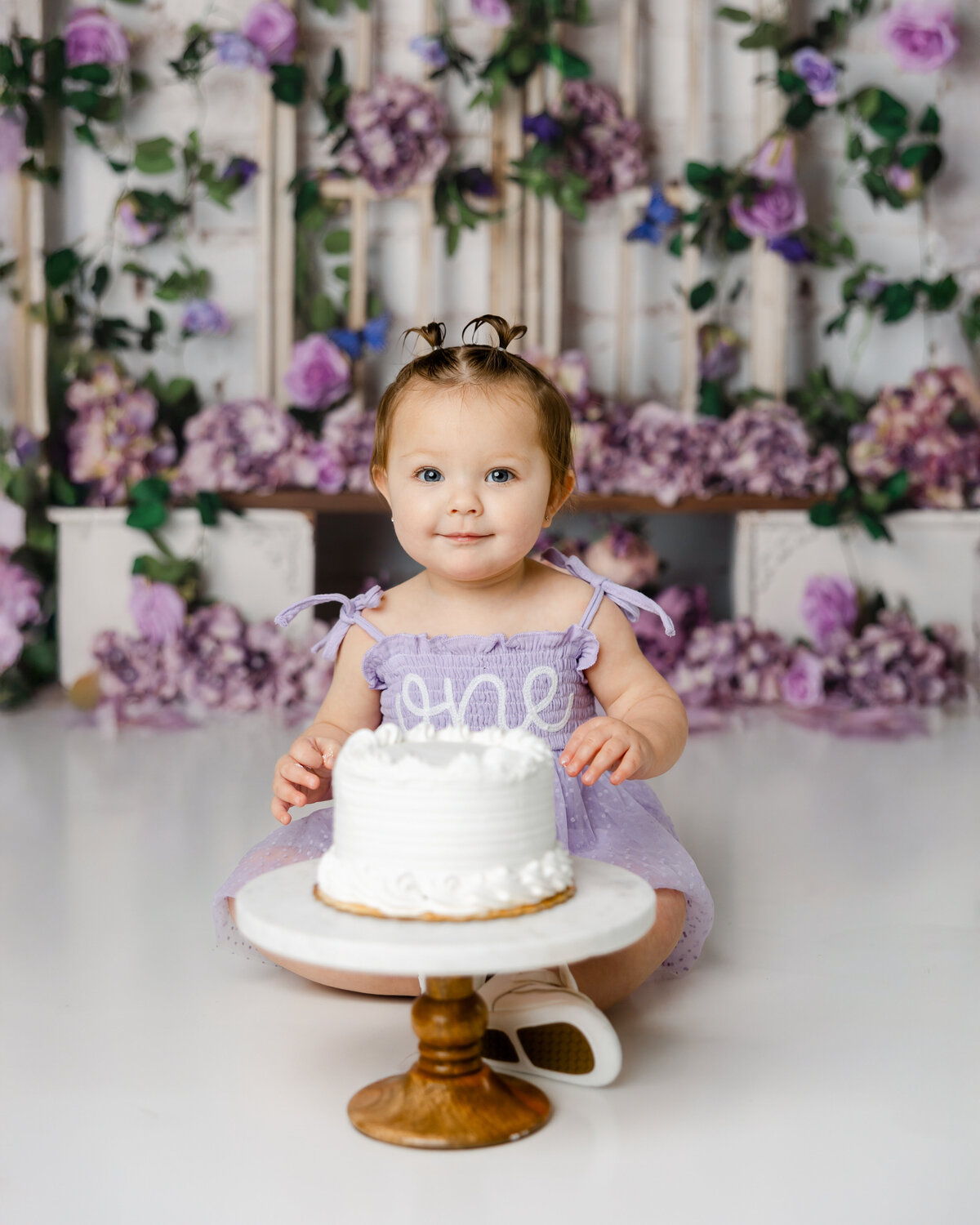 Baby in a lavender “one” dress sitting with a white cake in front of a floral purple backdrop. Photograph by Yucaipa cake smash photographer Kaitlyn Dawn Photography.