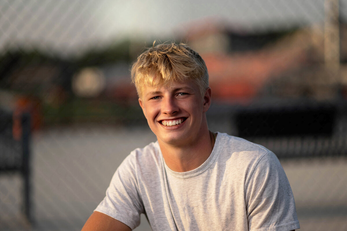 A blonde high school senior smiles while sitting in a white shirt at sunset