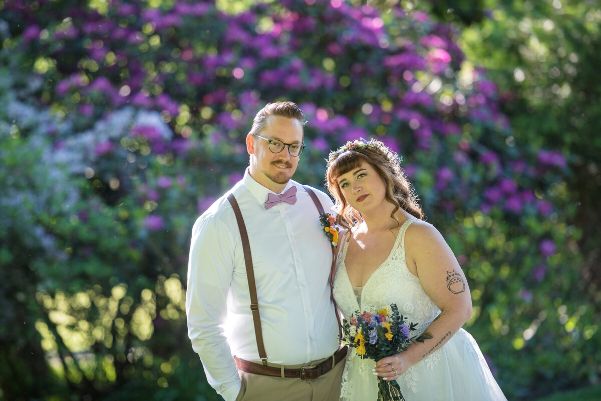 Bride with tattoo and groom in front of flowering bushes.