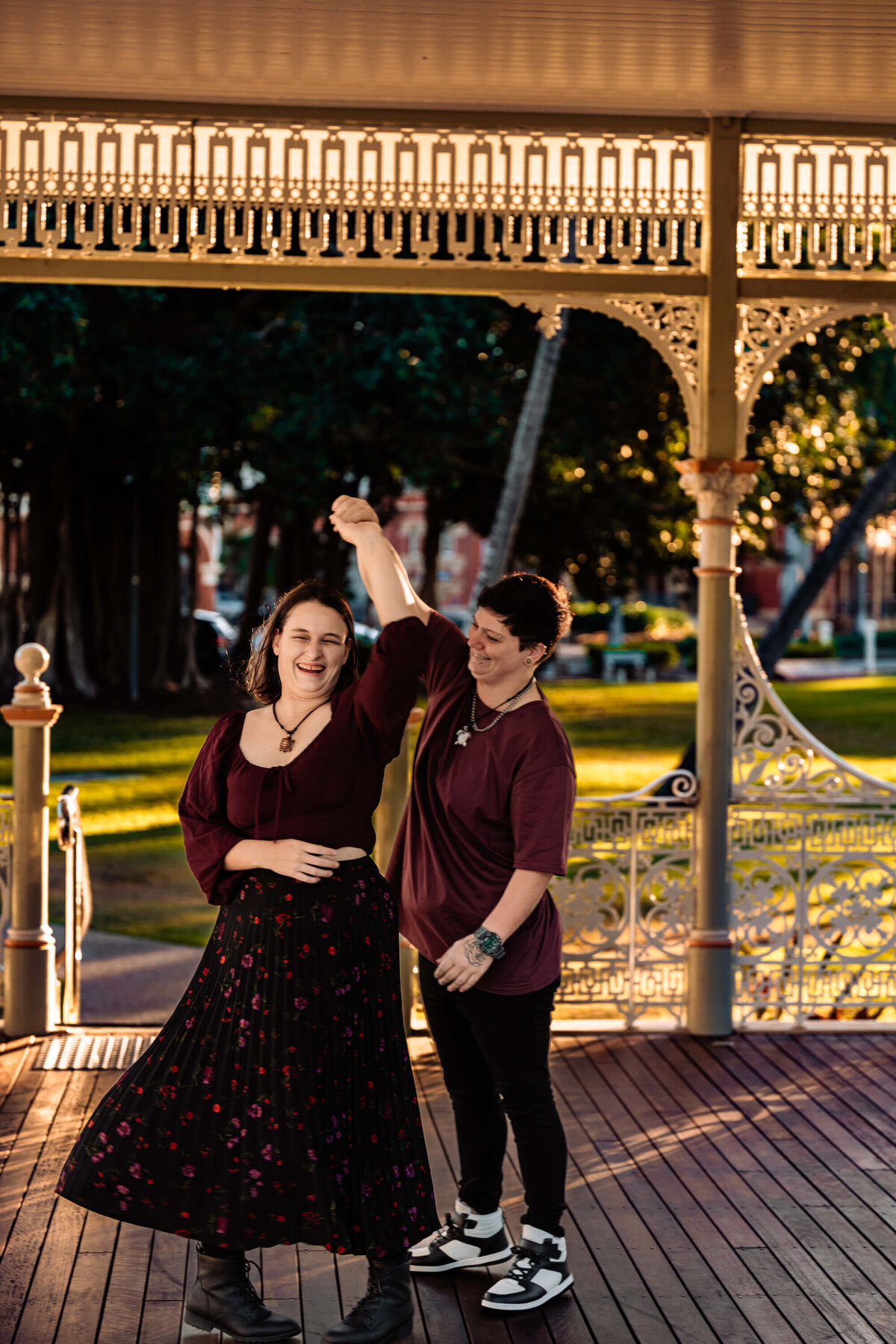 Two women joyfully dancing on a gazebo in a park, surrounded by greenery and a vibrant atmosphere. - photo by Jamie Simmons