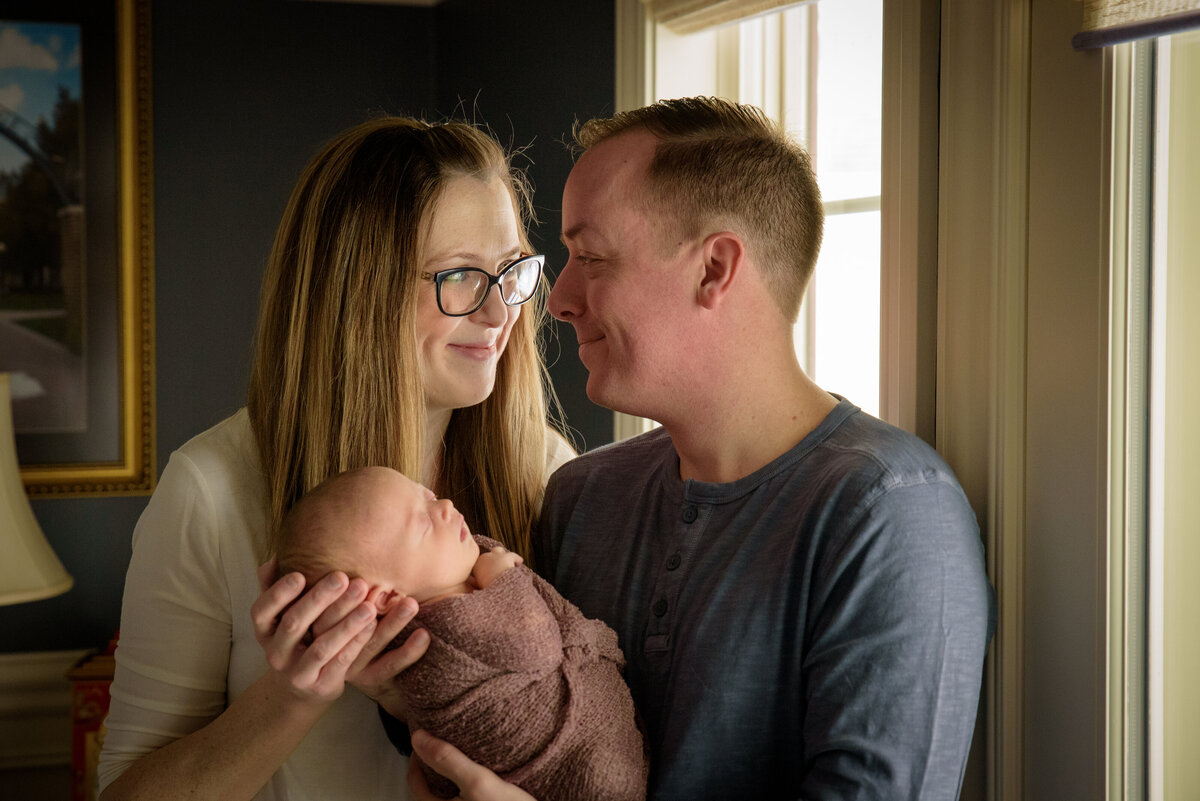 Mom & Dad are holding and adoring their new baby girl who is wearing a dusty rose wrap in front of a window in their home in Green Bay, Wisconsin.