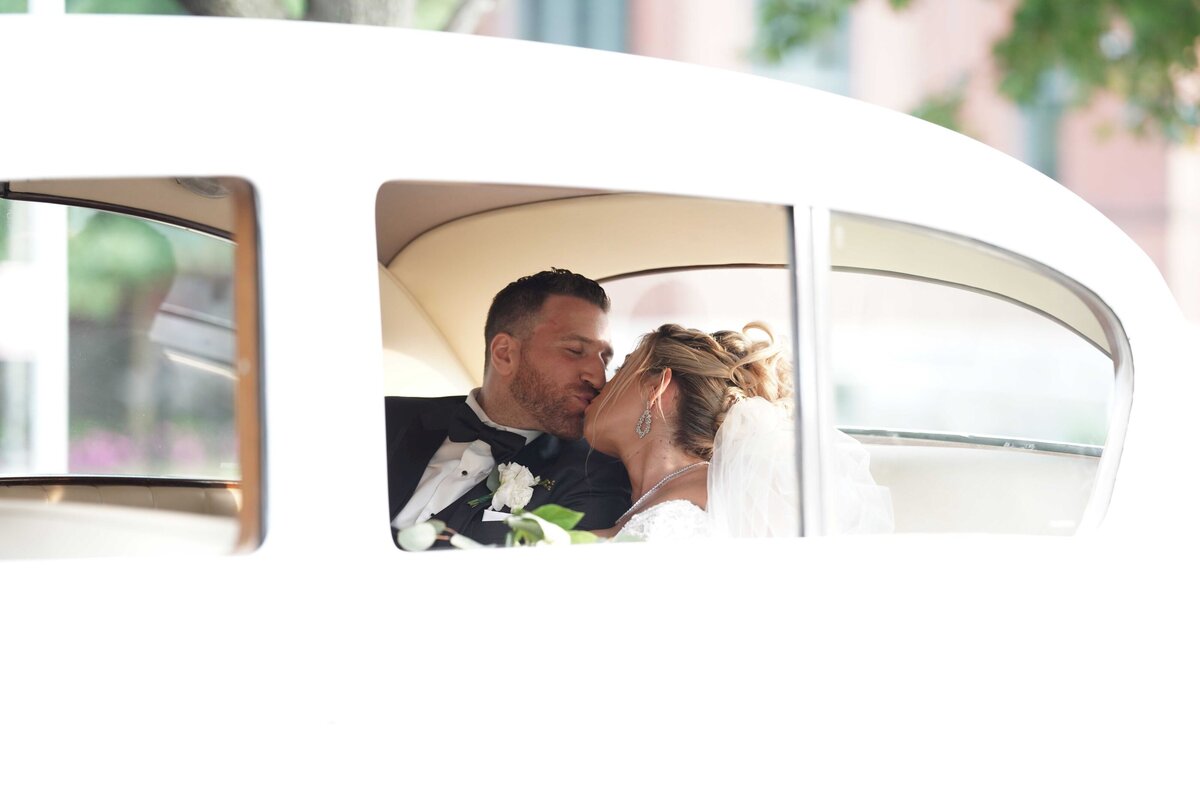 A couple is seen kissing tenderly in the backseat of a car, creating an intimate and romantic moment. The image highlights their closeness and affection, with the soft, cozy interior of the car setting a warm and private atmosphere.