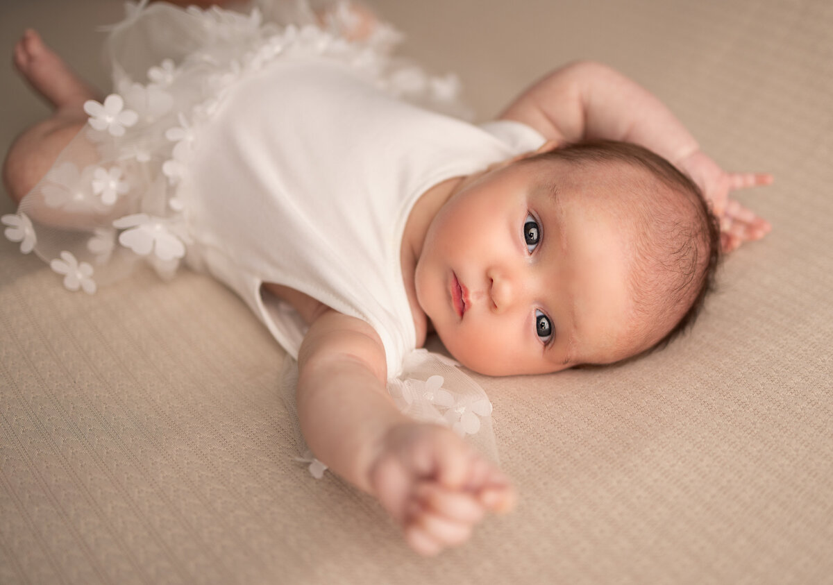 Awake newborn baby girl in a white floral outfit stretching her arm during a bright and natural light studio session in NJ
