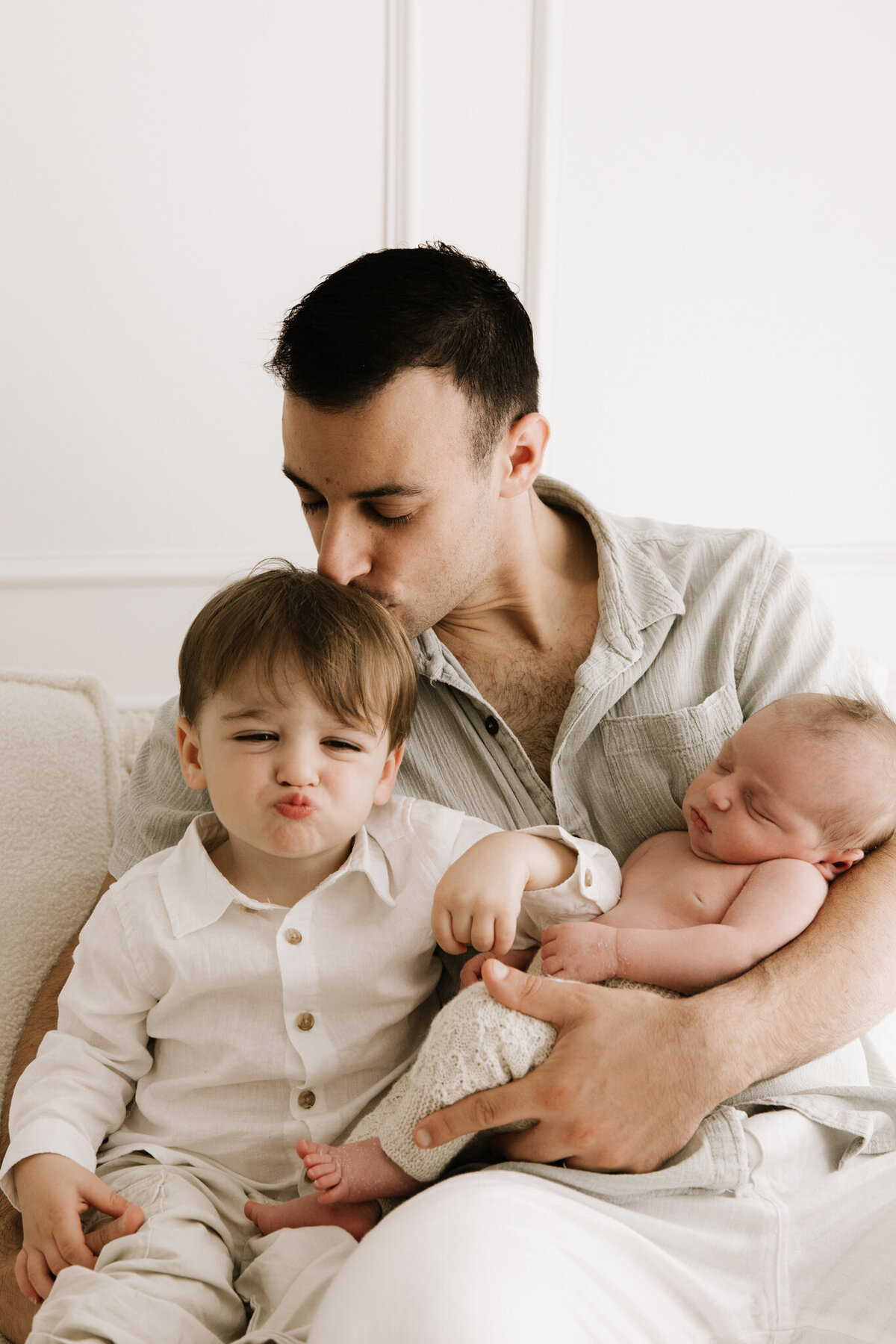 A father sits on a cream couch holding his two children, gently kissing his toddler on the head while the toddler makes a playful face. The newborn sleeps peacefully in his arms, wrapped in soft neutral tones. A sweet and candid family moment captured in studio lighting.