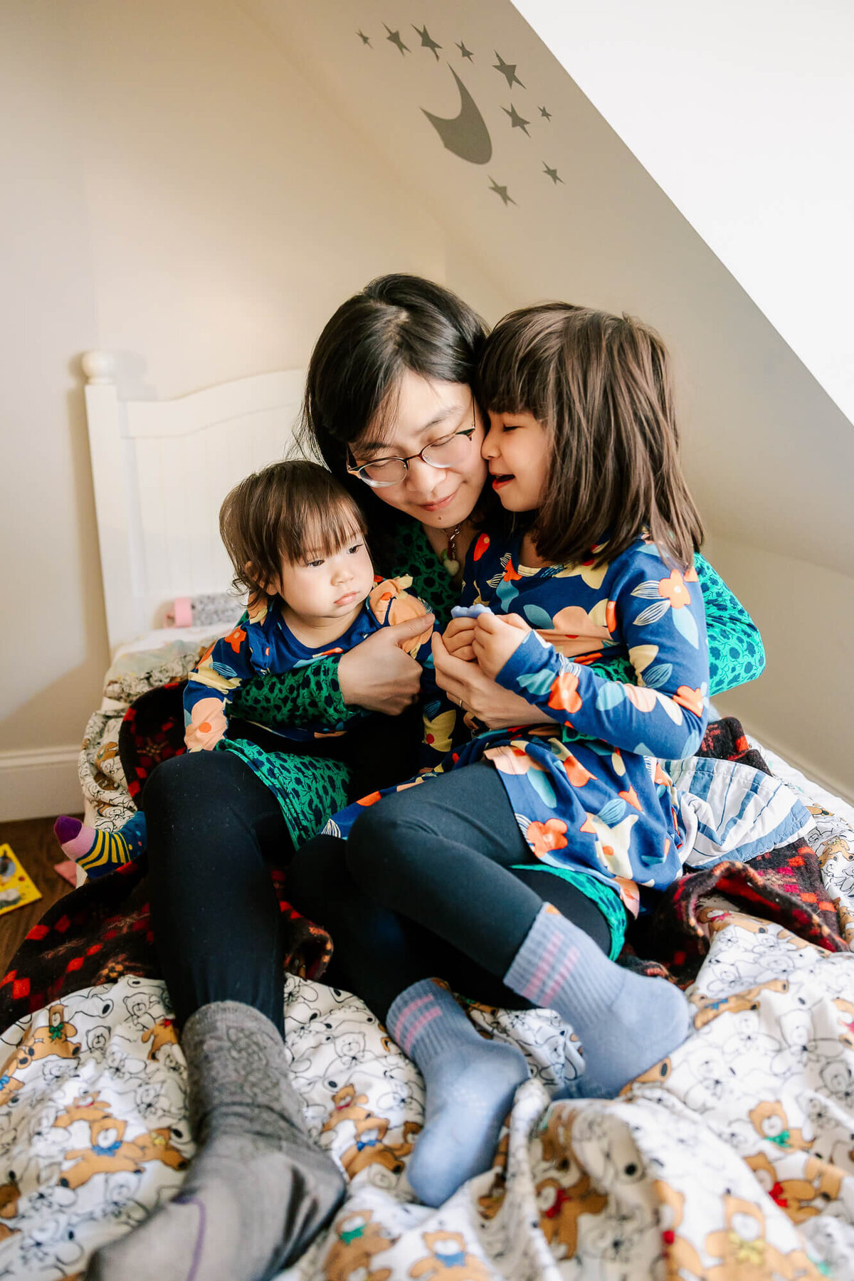Mother holds her two daughters close, sitting on a twin bed under a skylight in their Brookline home.