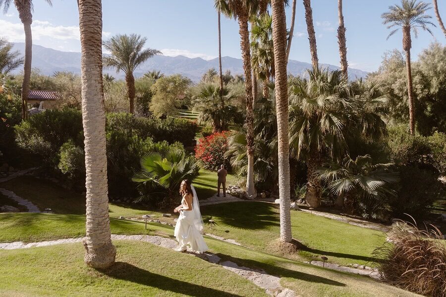 Bride walking down a rock path in the Oasis gardens while groom waits with his back turned.