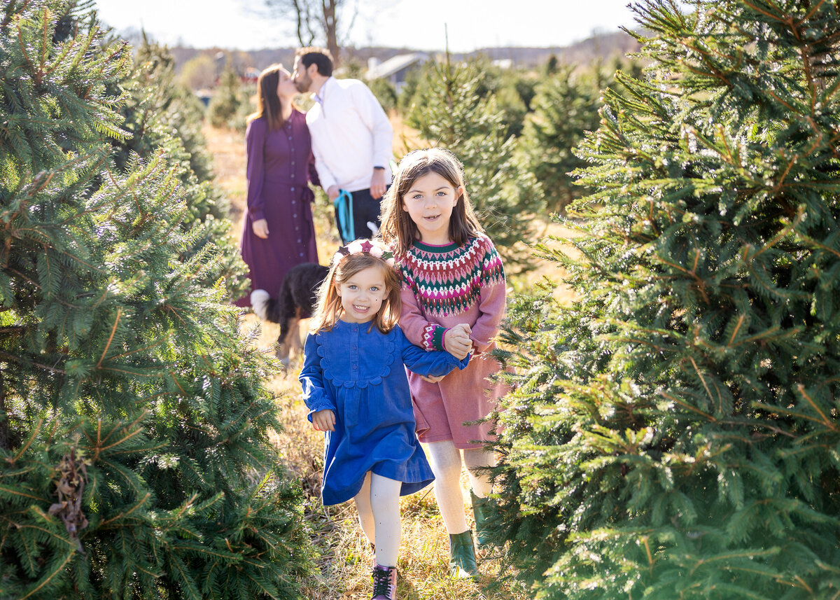 Siblings smile while mom and dad kiss in the background at a Christmas tree farm in Somerset County, NJ