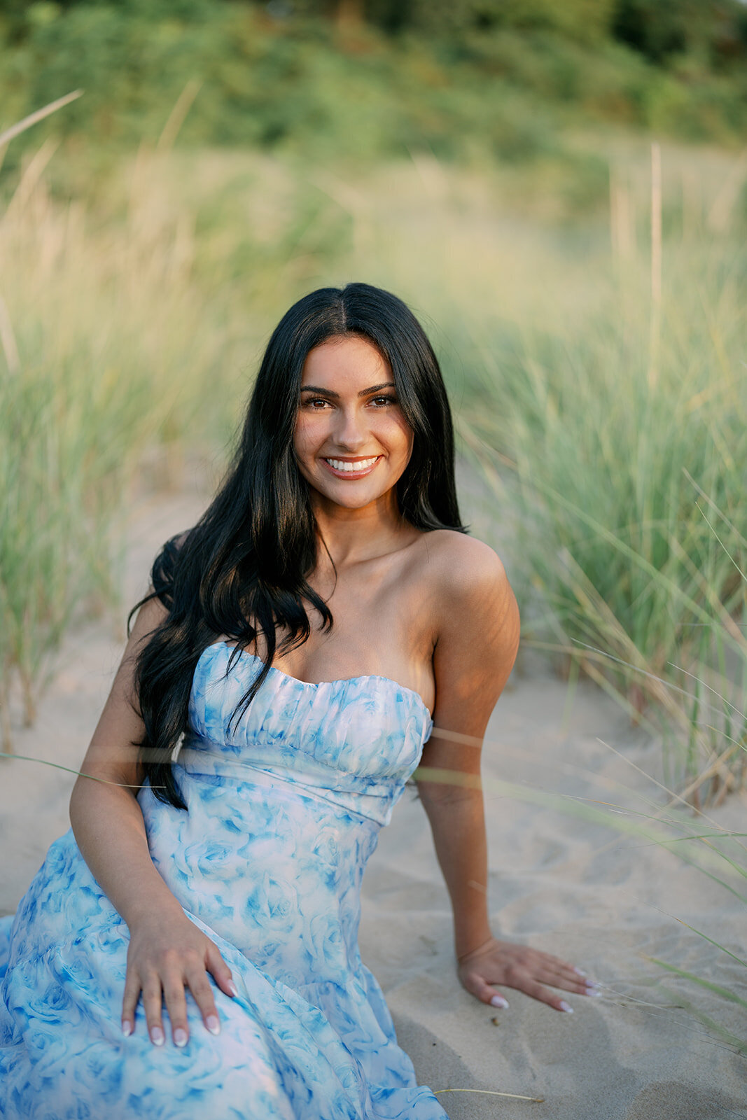 Senior girl sitting in the sand and dune grass wearing a blue dress during her Lake Michigan senior portraits.