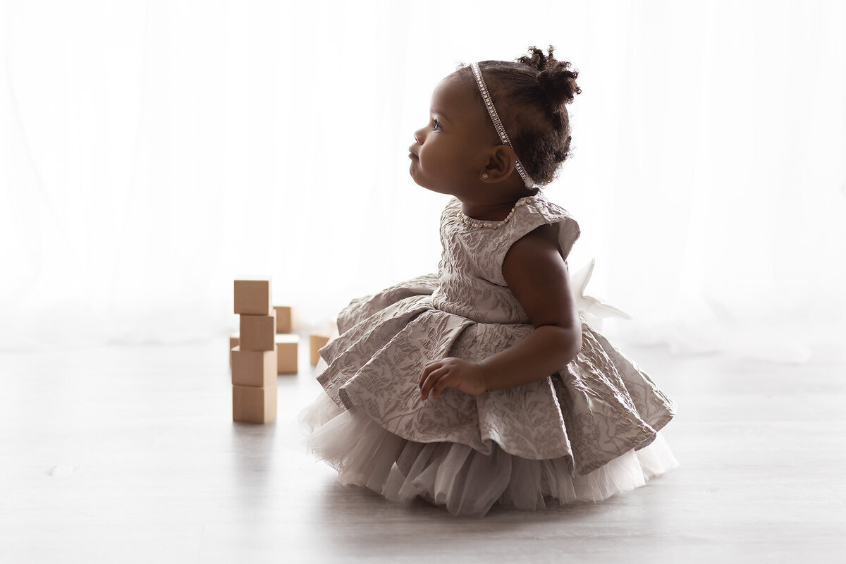 A toddler in a layered tulle dress and pearl headband gazes upward in soft backlight, sitting gracefully near wooden blocks during her Dallas milestone photographer session.