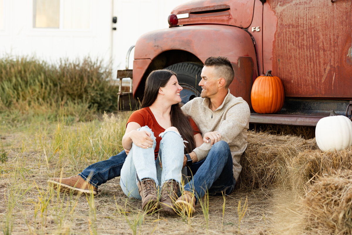 Husband and wife sit in the grass in front of a vintage red farm truck and smile at each other.