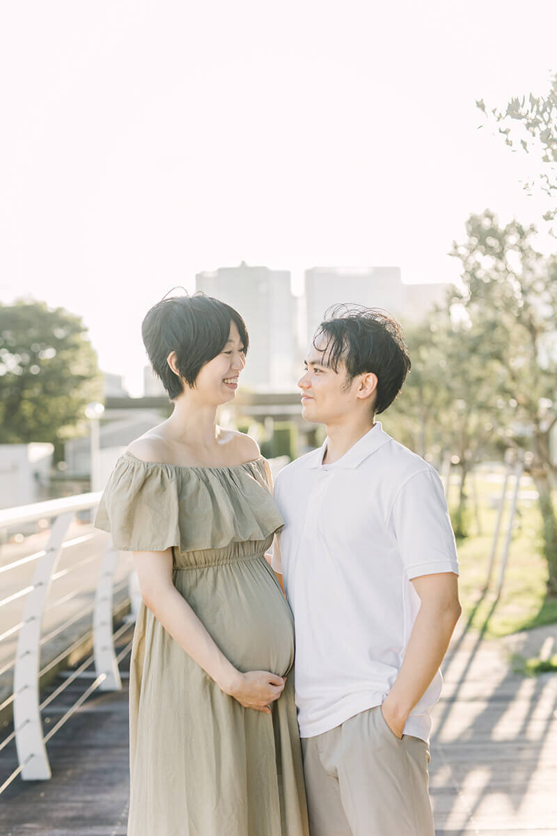 品川区で撮影。カーキベージュのワンピースを着た妊婦さんのママとパパが寄り添って目を合わせている、おしゃれで素敵なマタニティフォト。妊婦さんは手をおなかに添えて微笑んでいる。