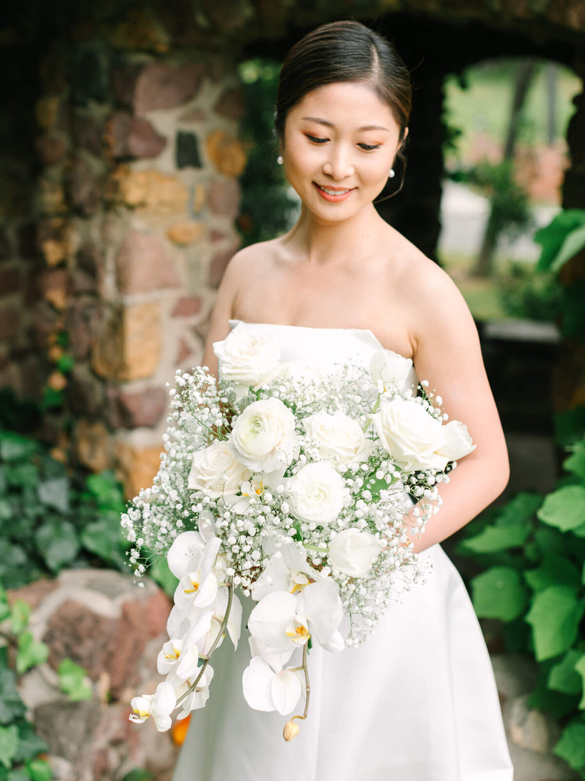 Bride in a strapless white gown holding a bouquet of white roses, orchids, and baby's breath. She smiles softly against a rustic stone wall and greenery.