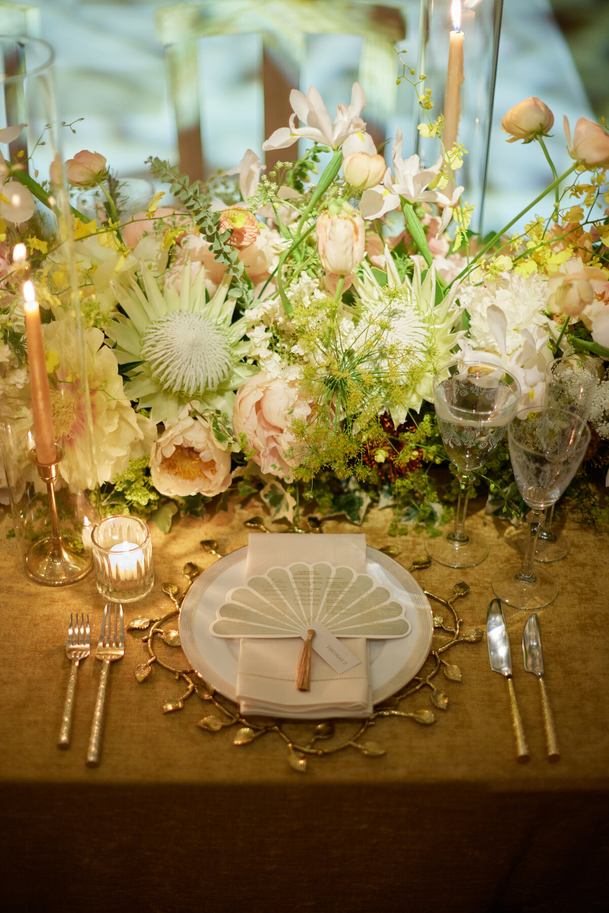 This beautifully arranged wedding table setting with an intricate floral centerpiece, featuring a variety of white, pink, and yellow flowers, was captured at the New York Public Library in New York City by photographer Shawn Connell. The table is set with elegant dinnerware, gold cutlery, and glassware, creating a sophisticated and romantic atmosphere, with Aaron Navak Films as Videographers capturing the special moments. A folded fan-shaped napkin resting on the dinner plate adds a touch of refinement to the overall presentation.