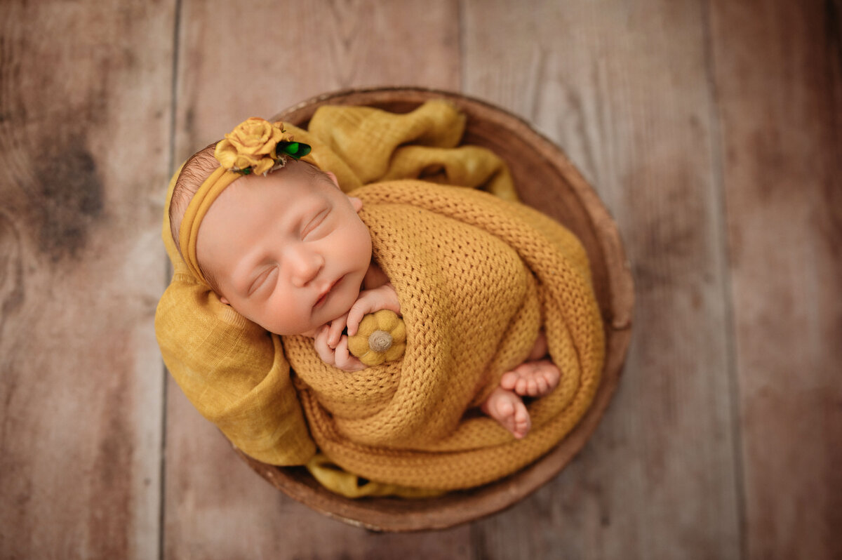 newborn baby girl wrapped in mustard yellow knit wrap with feet showing and holding a tiny felt yellow pumpkin  wearing floral headband on wooden backdrop