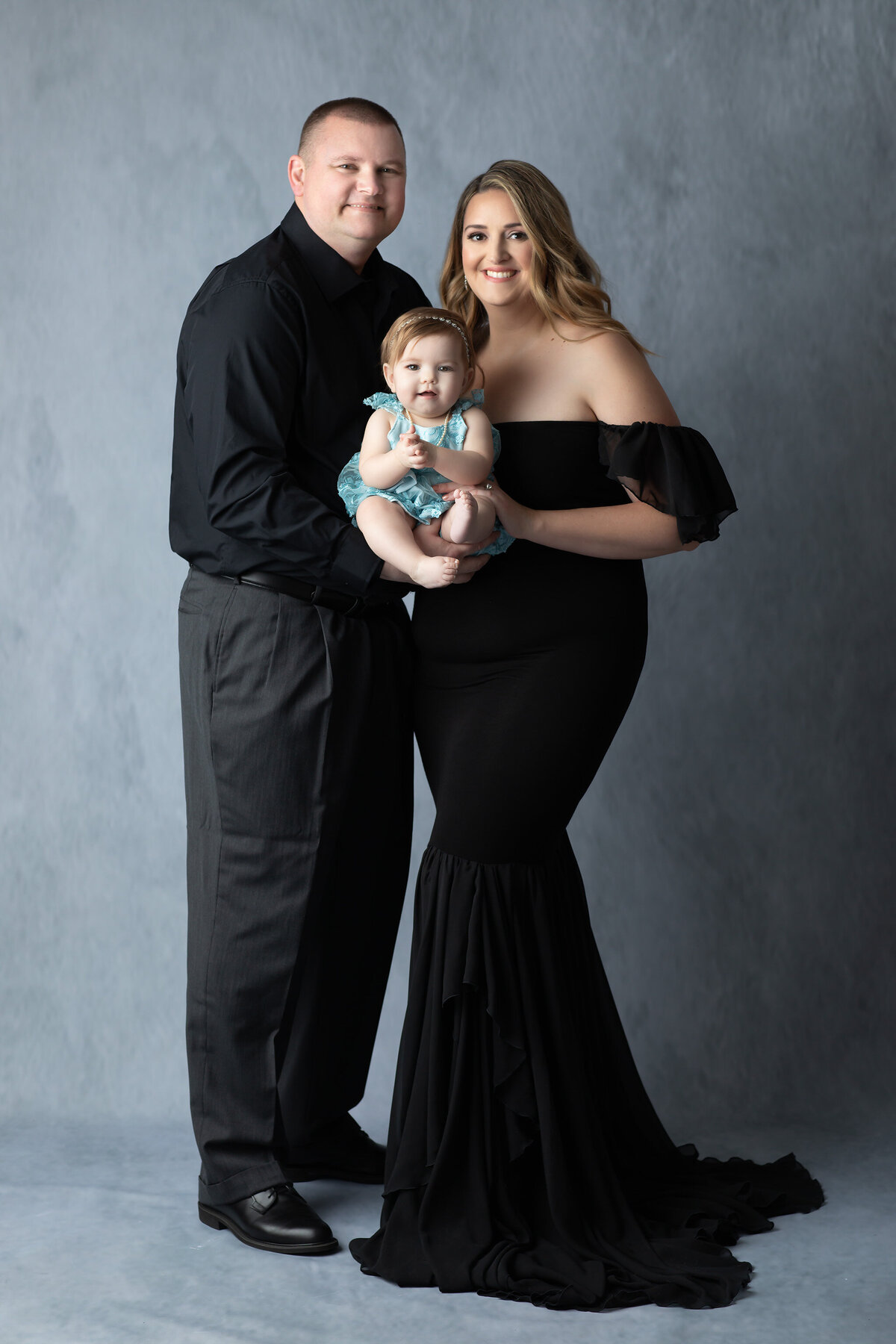 DFW milestone photographer captures a beautifully styled family portrait with mom in a flowing black gown, dad in formal attire, and their baby girl in a teal dress, all smiling warmly against a soft gray backdrop.